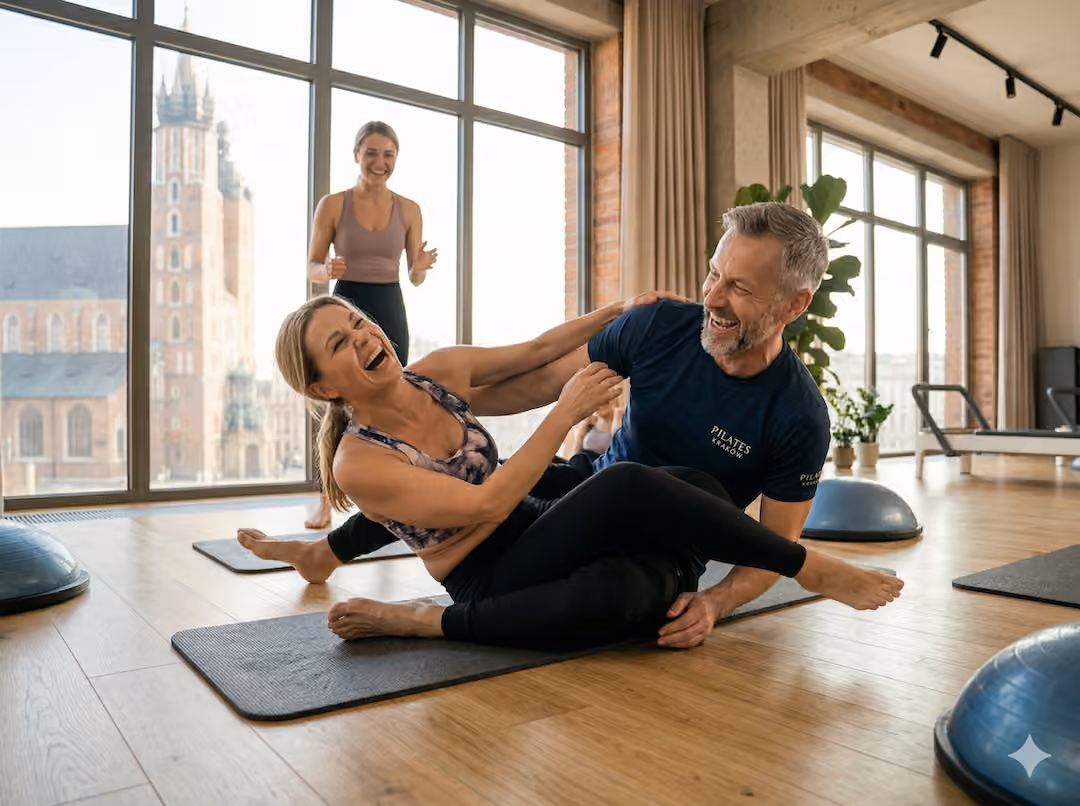Para po 50-tce w studiu pilates śmieje się podczas nieudanego ćwiczenia równoważnego, instruktor uśmiecha się w tle